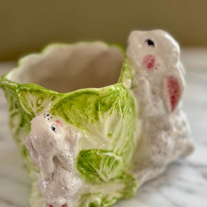 May include: A ceramic planter featuring a white bunny and two smaller bunnies. The planter is shaped like a lettuce head with green leaves. The bunnies have pink accents on their cheeks and ears. The planter is on a marble surface.