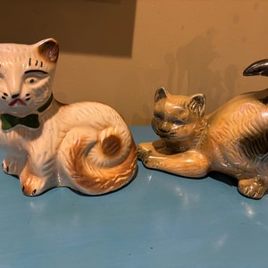 May include: Two ceramic cat figurines. One cat is white with brown spots and a green bow tie. The other cat is brown with black spots and a curled tail.