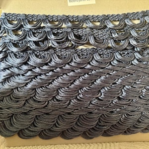 May include: A roll of gray decorative trim with a scalloped edge design. The trim is made of a silky material and is tightly wound on a cardboard spool. The trim is a muted gray color and appears to be new.
