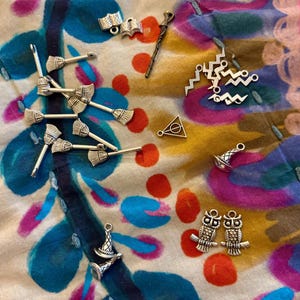 May include: A collection of silver-colored charms, including miniature brooms, open books, wizard hats, owls, a wand, and a triangle with a circle and line. The charms are scattered on a colorful fabric background with blue, pink, and yellow patterns.