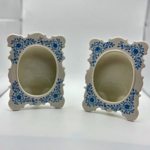 May include: Two off-white ceramic picture frames with oval openings. Each frame is decorated with blue floral patterns around the edges. The frames have a decorative, ornate shape.