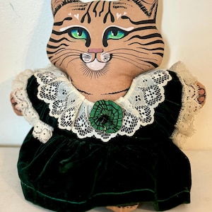 May include: A handmade plush cat doll wearing a green velvet dress with white lace trim and a green button. The cat has brown and black stripes, green eyes, and a painted smile.