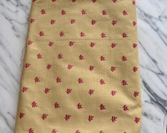 May include: Folded tan fabric with a repeating pattern of small, magenta-colored leaf-like designs. The fabric is folded neatly, showing the texture and pattern. The fabric is likely used for sewing or crafting.