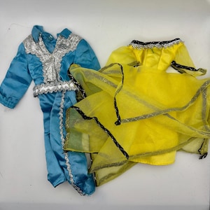 May include: Two doll outfits. One is a light blue satin-like jumpsuit with silver trim. The other is a yellow dress with black trim and a sheer skirt overlay. Both outfits are on a white surface.