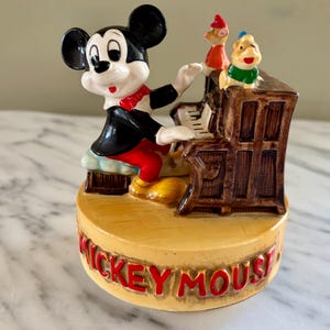 May include: A ceramic figurine featuring Mickey Mouse playing a piano. Mickey is dressed in a black jacket, red shorts, and a red bow tie. Two other cartoon characters are perched on top of the piano. The base of the figurine reads "Mickey Mouse" in red letters.