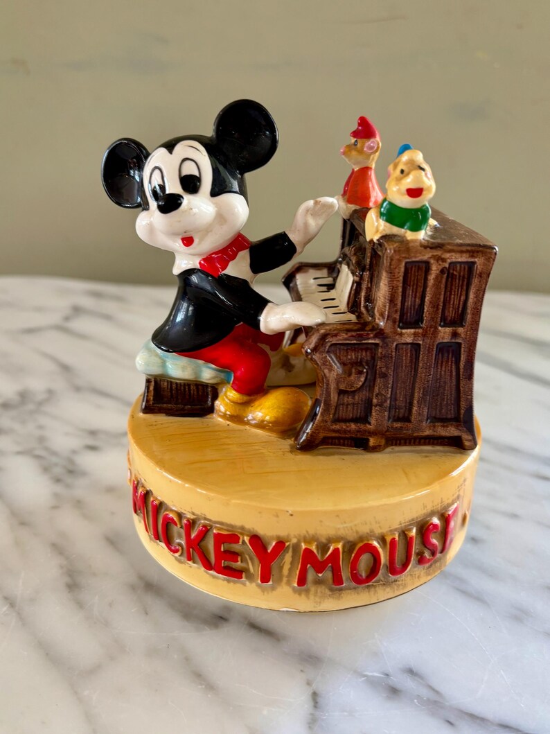May include: A ceramic figurine featuring Mickey Mouse playing a piano. Mickey is dressed in a black jacket, red shorts, and a red bow tie. Two other cartoon characters are perched on top of the piano. The base of the figurine reads "Mickey Mouse" in red letters.
