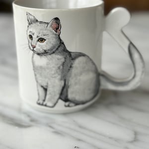May include: White ceramic mug with a gray cat illustration. The cat's tail is the handle of the mug.