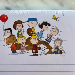 May include: A white notepad featuring a colorful illustration of the Peanuts gang. The image includes Charlie Brown, Snoopy, and other characters. A red balloon is visible in the upper left corner. The notepad has blue horizontal lines.