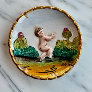 May include: A small, round ceramic dish with a gold rim and a raised design of a cherub in a garden setting. The cherub is painted in shades of pink and white, and the garden is painted in shades of green, yellow, and brown.