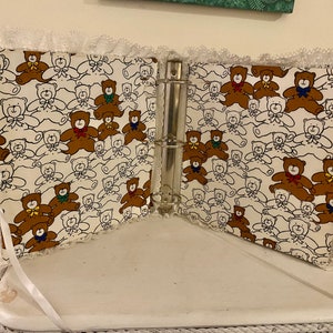 May include: A white binder with a lace trim and a teddy bear pattern. The binder is open and the rings are visible. The teddy bears are brown, white, and have different colored bows.