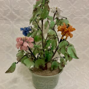 May include: A decorative glass flower arrangement in a green ceramic pot. The arrangement features colorful glass flowers in shades of pink, orange, blue, and white, with green glass leaves. The pot has a textured design.