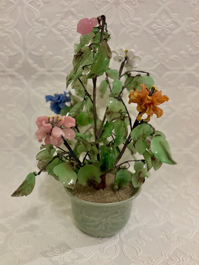 May include: A decorative glass flower arrangement in a green ceramic pot. The arrangement features colorful glass flowers in shades of pink, orange, blue, and white, with green glass leaves. The pot has a textured design.