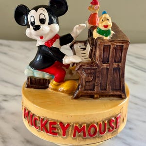 May include: A vintage Mickey Mouse figurine playing a piano. Mickey is wearing a black jacket, red pants, and a red bow tie. The piano is brown, and there are two other cartoon characters on top. The base is tan with "MICKEY MOUSE" in red.