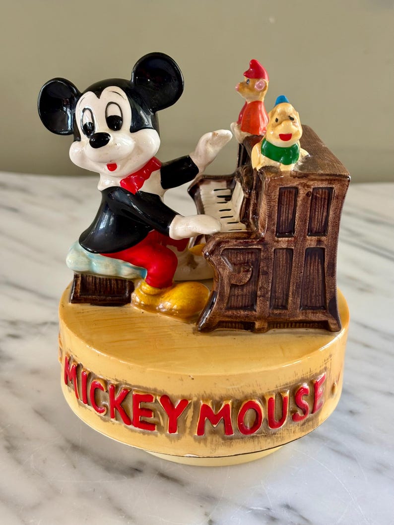 May include: A vintage Mickey Mouse figurine playing a piano. Mickey is wearing a black jacket, red pants, and a red bow tie. The piano is brown, and there are two other cartoon characters on top. The base is tan with "MICKEY MOUSE" in red.