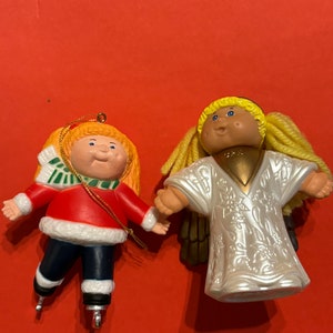 May include: Two Cabbage Patch Kids Christmas ornaments. One is a girl wearing a red and white outfit and ice skates. The other is an angel with a white dress and gold wings.