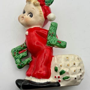May include: A vintage ceramic Christmas figurine of a child in a red outfit, holding a green wreath and a green stocking. The figurine has a white basket with gold accents and black shoes. A festive holiday decoration.