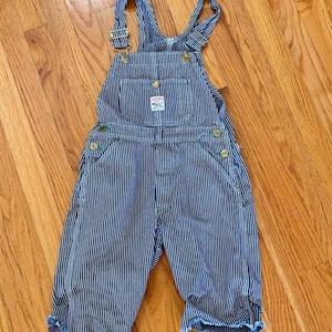 May include: Blue and white hickory striped denim overalls featuring adjustable shoulder straps with metal buckles, a front bib pocket with a rectangular patch label, side button closures, and frayed, raw-edge hems at the bottom of the pant legs.