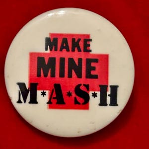 May include: A white button with a red cross and the text "MAKE MINE M*A*S*H" in black.