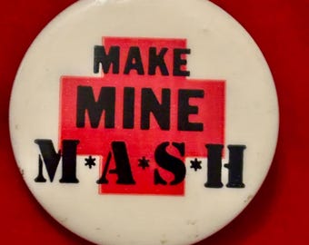 May include: A white button with a red cross and the text "MAKE MINE M*A*S*H" in black.