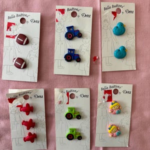 May include: A collection of Belle Buttons by Dritz, featuring various designs. The buttons include footballs, tractors, ducks, airplanes, and baby dolls. Each set is attached to a white card with a drawing of children.