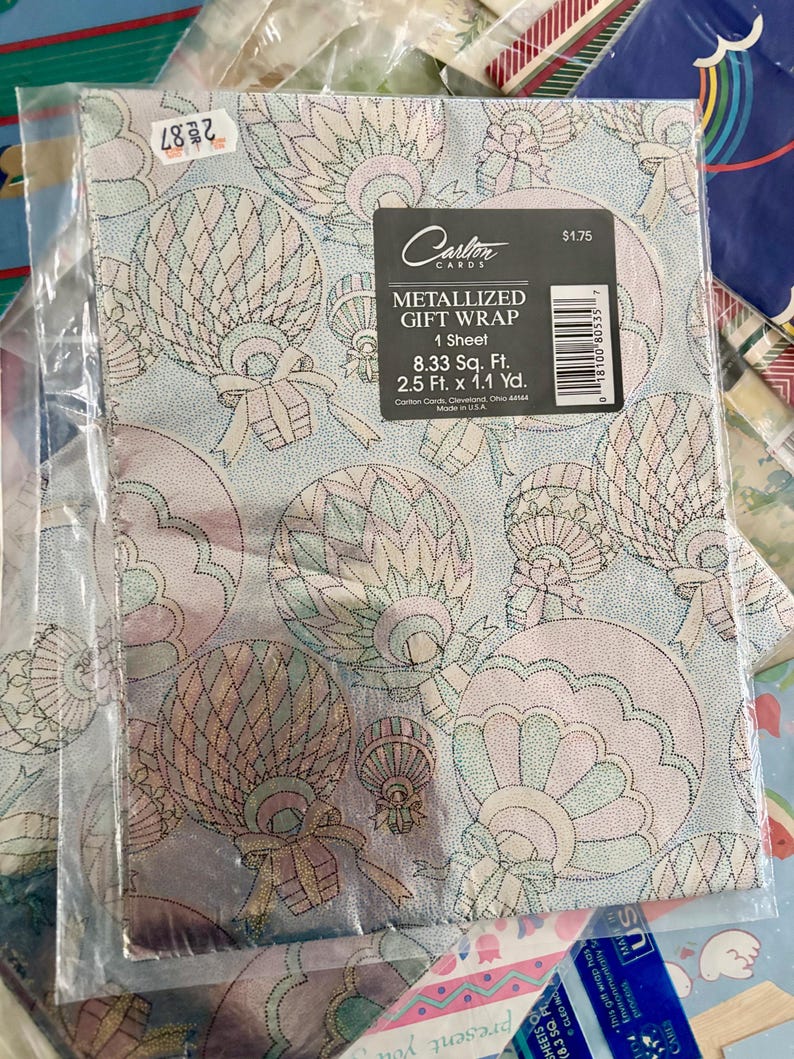 Peut inclure: Un rouleau de papier cadeau m&eacute;tallis&eacute; avec un fond bleu et un motif de montgolfi&egrave;res roses et vertes. Le papier cadeau mesure 8,33 pieds carr&eacute;s et 2,5 pieds sur 1,1 yard. Le texte "Carlton Cards" et "Metallized Gift Wrap" est imprim&eacute; sur le papier cadeau.