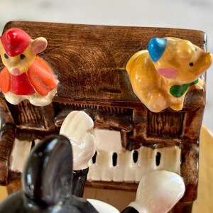 May include: A ceramic music box featuring a piano with a brown finish. The piano has white keys and is decorated with cartoon animal figurines, including a mouse in a red outfit and a pig with a blue ear.