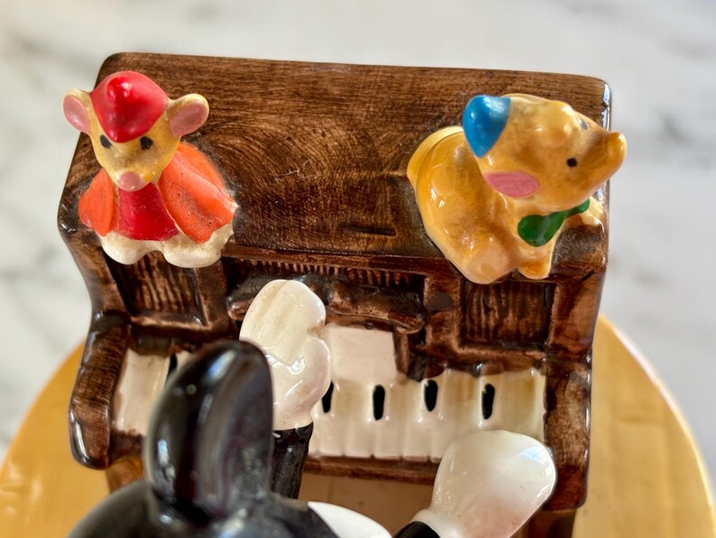 May include: A ceramic music box featuring a piano with a brown finish. The piano has white keys and is decorated with cartoon animal figurines, including a mouse in a red outfit and a pig with a blue ear.
