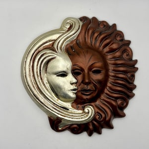 May include: A decorative wall hanging featuring a sun and moon design. The moon is silver with a face, and the sun is brown with a face. The two figures are side by side, with the moon on the left and the sun on the right. The wall hanging is made of a hard material.