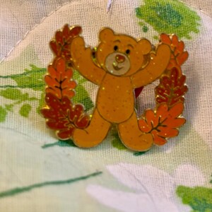 May include: A gold-colored bear pin with outstretched arms, surrounded by a wreath of orange and red autumn leaves. The bear has a glittery finish and a red heart detail. The pin is set against a floral patterned background.