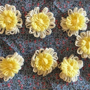 May include: A collection of handmade yarn flowers with yellow centers and white petals. The flowers are arranged on a blue floral patterned fabric, creating a cheerful and decorative display. These could be used for crafting or embellishments.