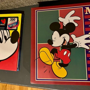 May include: Two photo albums featuring Mickey Mouse. One album has a large image of Mickey's face on a yellow, red, and blue background. The other album has a full image of Mickey Mouse with the word "MICKEY" vertically on the side.
