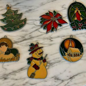 May include: Set of ten stained glass Christmas ornaments featuring a variety of holiday themes, including a Christmas tree, a poinsettia, a snowman, a church, a lamb, and a candle. The ornaments are made of glass and have a metallic finish.