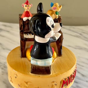 May include: A vintage Mickey Mouse figurine playing a piano, with two other cartoon characters on top. The figurine is on a yellow base with red lettering. Mickey is wearing a black jacket, red shorts, and yellow shoes.