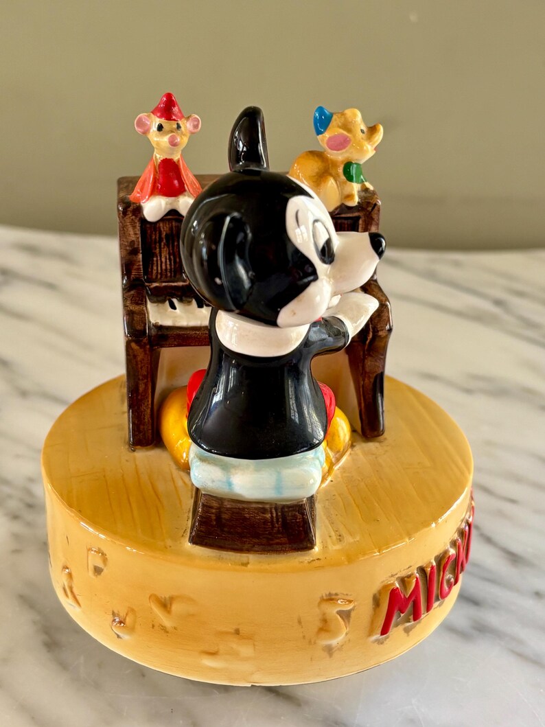 May include: A vintage Mickey Mouse figurine playing a piano, with two other cartoon characters on top. The figurine is on a yellow base with red lettering. Mickey is wearing a black jacket, red shorts, and yellow shoes.