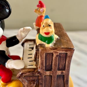 May include: A vintage figurine featuring Mickey Mouse playing a brown piano, with two other cartoon characters standing on top. The figurine has a glossy finish and is painted in bright colors.