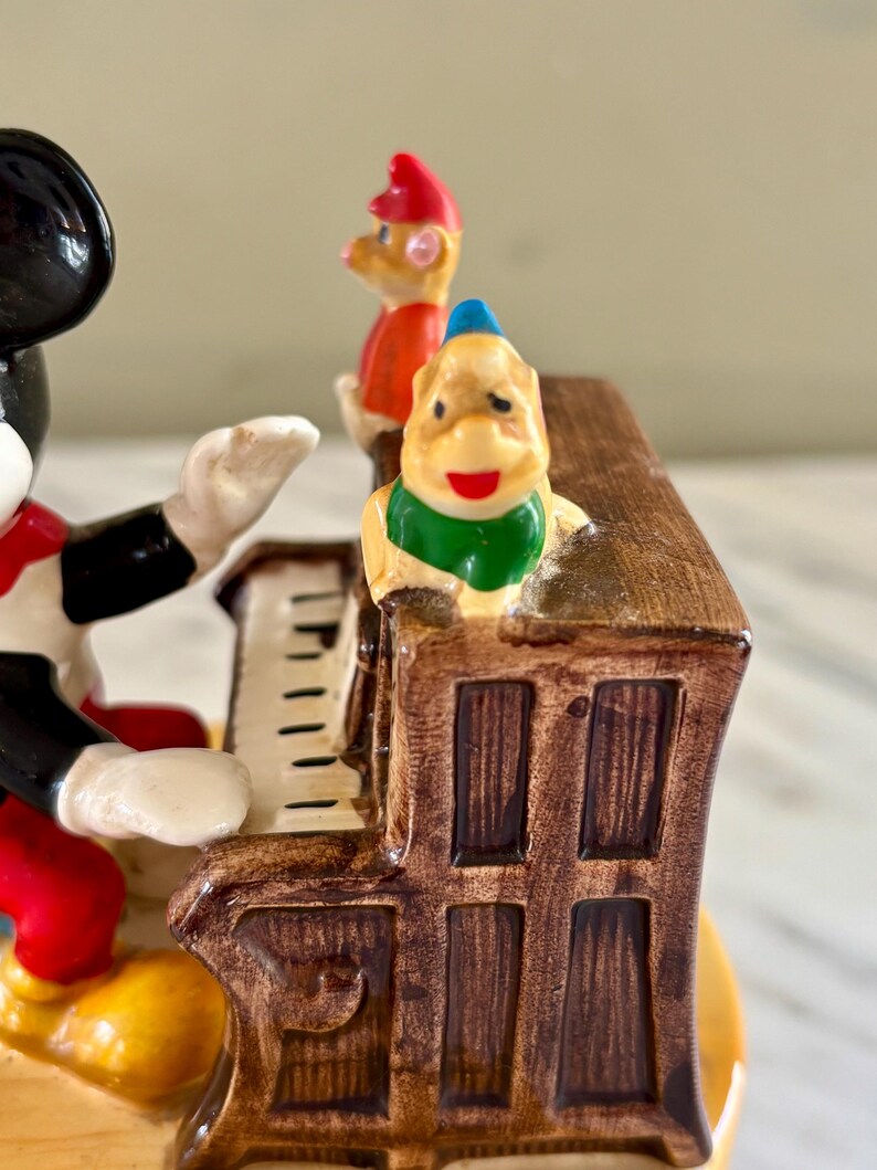 May include: A vintage figurine featuring Mickey Mouse playing a brown piano, with two other cartoon characters standing on top. The figurine has a glossy finish and is painted in bright colors.