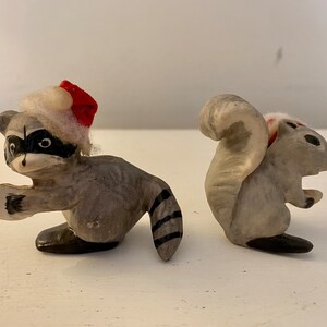 May include: Two small, hand-painted figurines of a raccoon and a squirrel. The raccoon is wearing a red and white Santa hat and has a black and white striped tail. The squirrel has a white and red tail and is sitting up with its tail curled around its body.