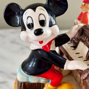 May include: A ceramic figurine of Mickey Mouse playing a piano. Mickey is wearing a black tuxedo, red pants, and yellow shoes. The piano is brown with white keys. A small figure of Pinocchio is visible on the piano.