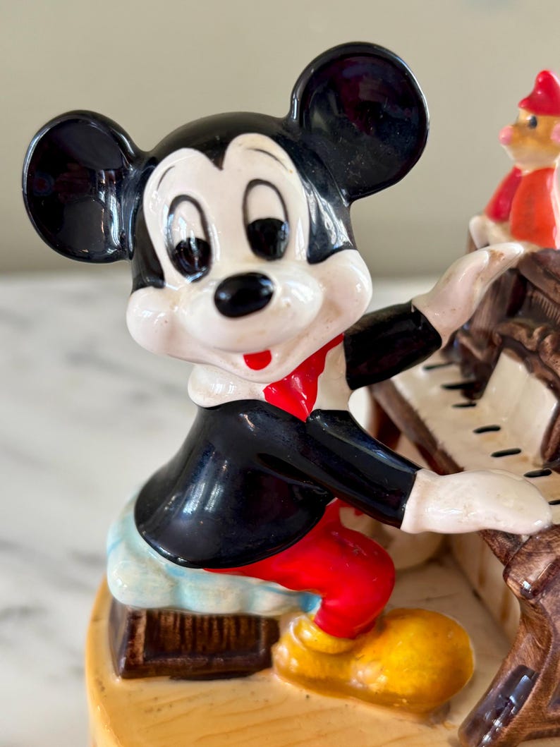 May include: A ceramic figurine of Mickey Mouse playing a piano. Mickey is wearing a black tuxedo, red pants, and yellow shoes. The piano is brown with white keys. A small figure of Pinocchio is visible on the piano.