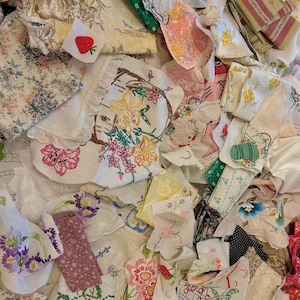 May include: A collection of vintage fabric scraps, featuring floral patterns, embroidered designs, and various textures. The color palette includes pinks, greens, yellows, and whites, with a small embroidered strawberry patch.