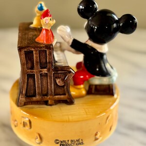 May include: A vintage Mickey Mouse figurine playing a piano, with a small figure on top of the piano. The figurine is on a yellow base with musical notes and the text "WALT DISNEY PRODUCTIONS, JAPAN". Mickey is wearing a black tuxedo and red pants.