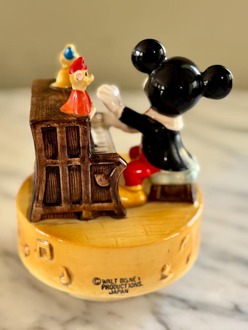 May include: A vintage Mickey Mouse figurine playing a piano, with a small figure on top of the piano. The figurine is on a yellow base with musical notes and the text "WALT DISNEY PRODUCTIONS, JAPAN". Mickey is wearing a black tuxedo and red pants.
