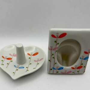 May include: A white ceramic ring holder in a heart shape and a small picture frame. Both items are decorated with a floral design in orange, pink, and blue. The ring holder has a central post.