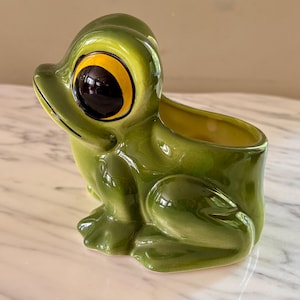 May include: A glossy green ceramic frog planter featuring a large, prominent eye with a dark pupil and a bright yellow iris. The frog is depicted in a seated position with a hollow opening on its back, finished with a smooth, reflective glaze.