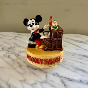 May include: A vintage Mickey Mouse figurine playing a piano. The figurine features Mickey in a black suit with a red bow tie, and two other characters. The base reads "Mickey Mouse" in red letters.