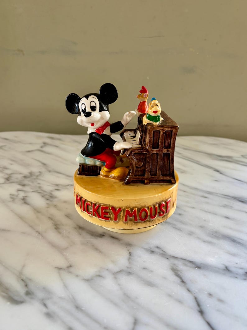 May include: A vintage Mickey Mouse figurine playing a piano. The figurine features Mickey in a black suit with a red bow tie, and two other characters. The base reads "Mickey Mouse" in red letters.