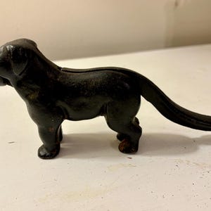 May include: A black cast iron dog-shaped nutcracker. The dog is standing with its tail extended. The nutcracker has a detailed design and a rustic appearance. The dog's mouth is open, ready to crack nuts.