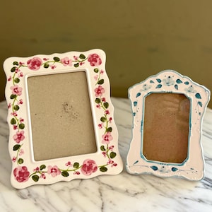 May include: Two decorative photo frames. One is white with pink roses and green leaves. The other is white with blue floral accents. Both frames have a rectangular opening for a photo.