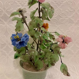 May include: A decorative glass flower arrangement in a light green pot. The arrangement features blue, pink, white, and orange flowers with green leaves. A pink fruit is at the top of the arrangement.