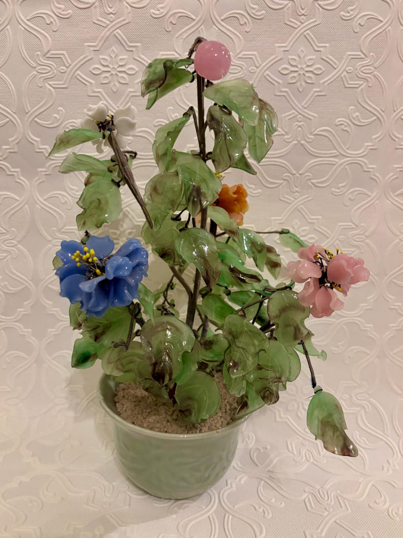 May include: A decorative glass flower arrangement in a light green pot. The arrangement features blue, pink, white, and orange flowers with green leaves. A pink fruit is at the top of the arrangement.